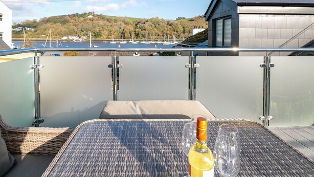 Uma varanda com uma mesa, uma garrafa de vinho e vista para barcos e colinas.