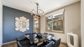 Un comedor con una mesa de vidrio, seis sillas, una araña y un amplio ventanal con vistas a edificios.