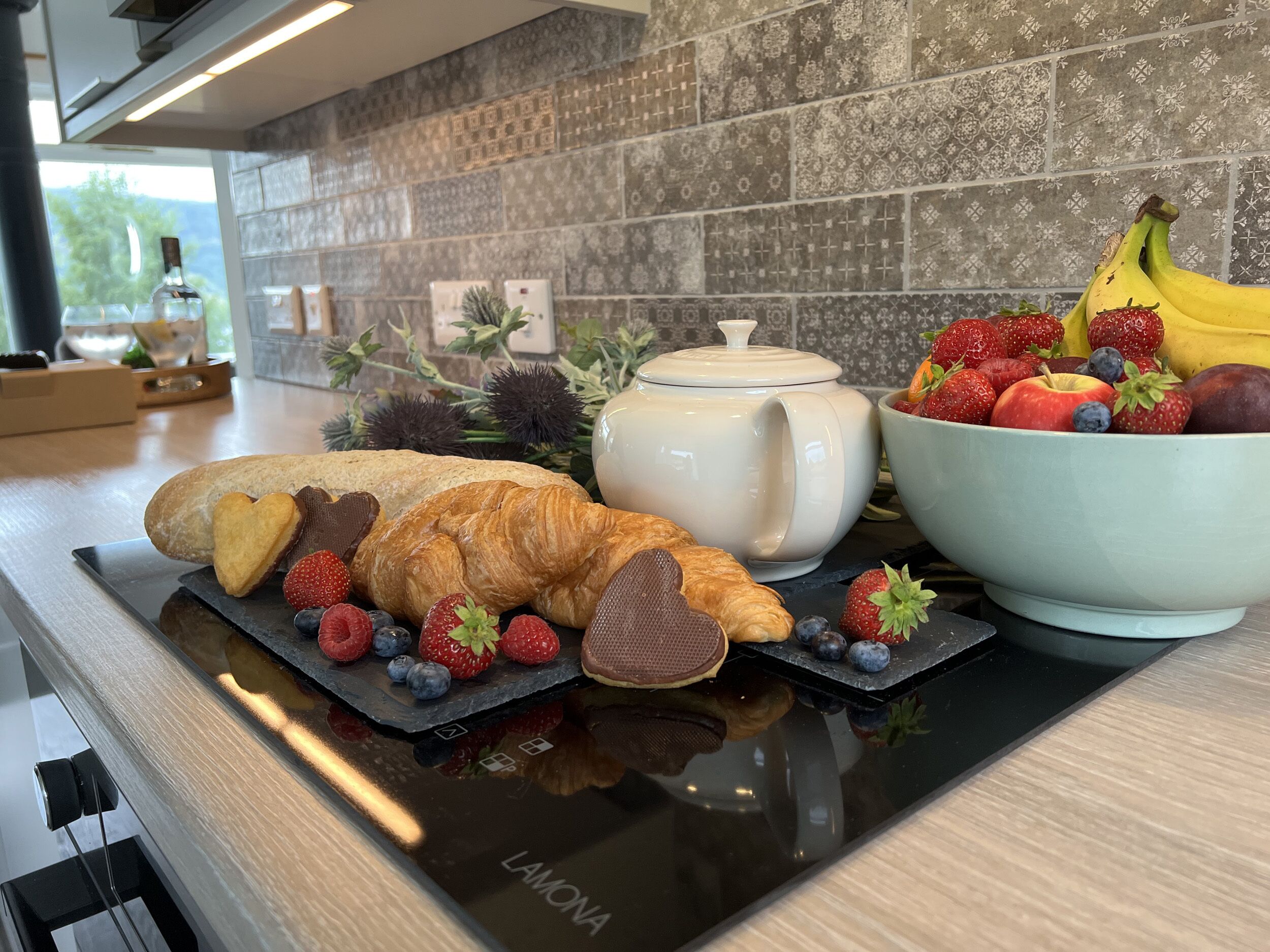A kitchen counter with a variety of food items including bread, pastries, and a bowl of fruit.