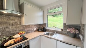 A modern kitchen with white cabinets, a stainless steel range hood, a toaster, and a window with a view of greenery. - Ciaran Cottage (Aberfeldy)
