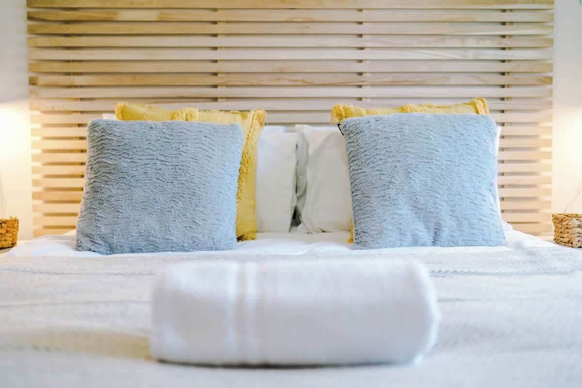 Een net opgemaakt bed met twee kussens, een wit en een blauw, en een opgerolde witte handdoek op het bed.