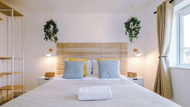 Een netjes ingerichte slaapkamer met een groot bed, twee nachtkastjes, een hangplant en een raam met gordijnen.