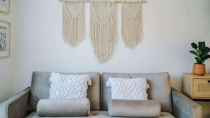 Een sofa in neutrale tinten met decoratieve kussens, een macramé wanddecoratie en een kamerplant op een houten bijzettafeltje.