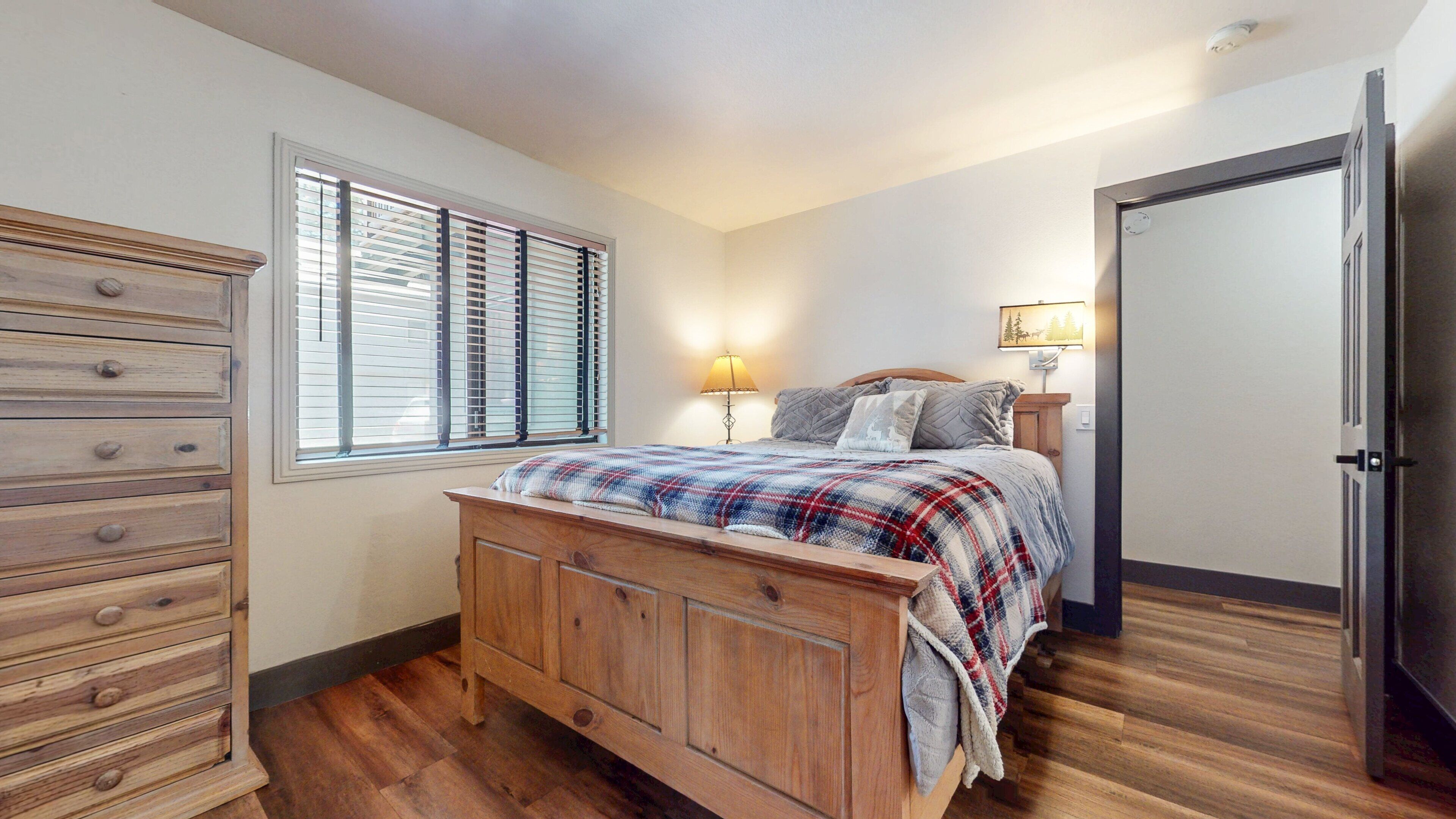 Un dormitorio con una cama de madera, un tocador, una ventana con persianas, una lámpara de noche y una puerta.
