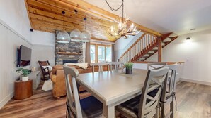 A spacious living area with a dining table, chairs, a fireplace, a staircase, and a TV mounted on the wall.