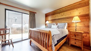 A wooden-framed bed with a headboard, two bedside tables with lamps, a wooden chair, and a large sliding glass door leading to an outdoor area.