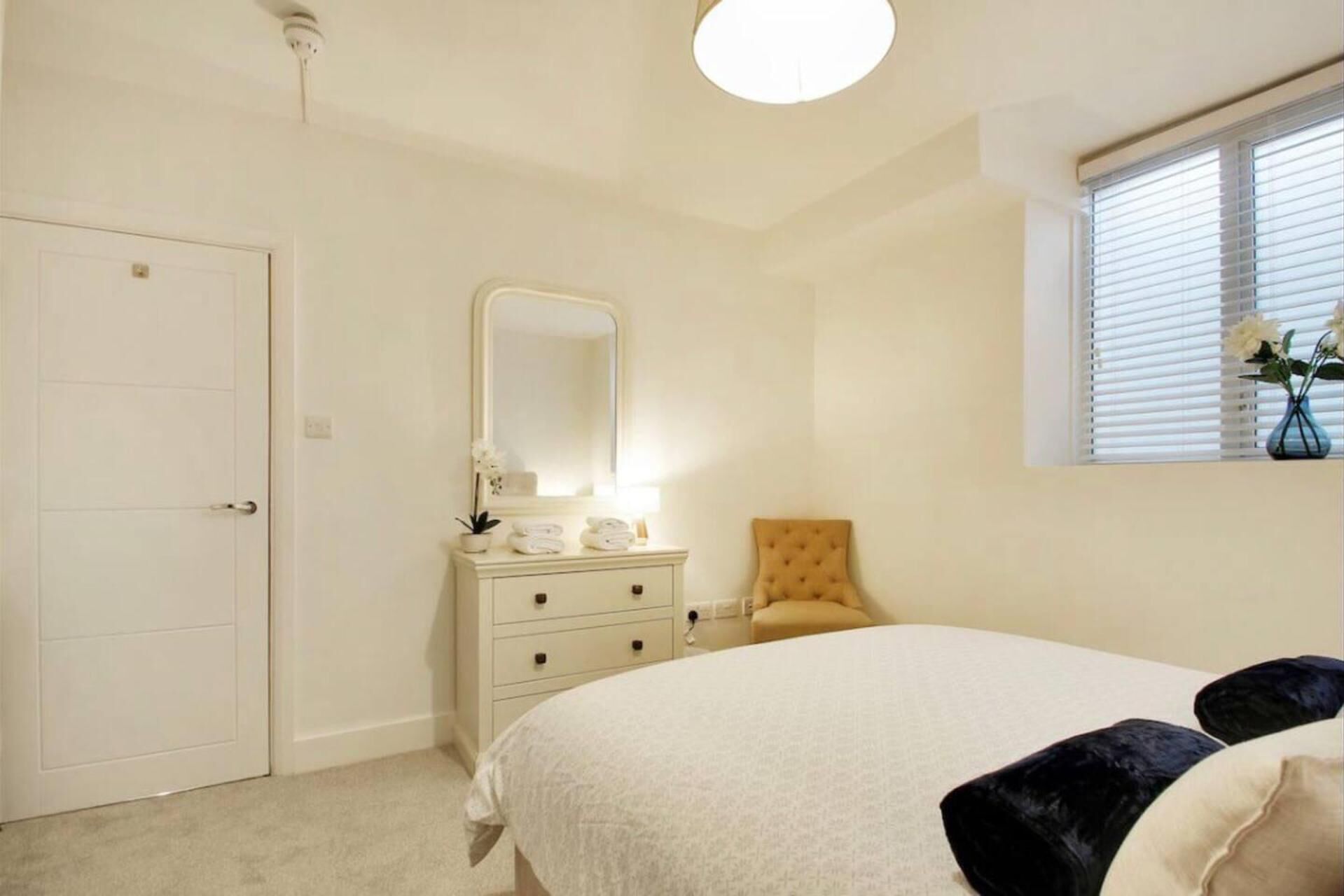 Een slaapkamer met een bed, een kaptafel met spiegel, een raam met jaloezieën en een deur.
