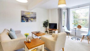 A modern living room with a sofa, coffee table, and TV stand. There is a window with curtains and a view of greenery outside. - Stylish 1-bed Apartment - Heart of Tunbridge Wells (Royal Tunbridge Wells)