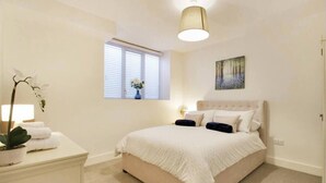 A bedroom with a bed, a nightstand, a lamp, a window with blinds, and a painting on the wall. - Stylish 1-bed Apartment - Heart of Tunbridge Wells (Royal Tunbridge Wells)