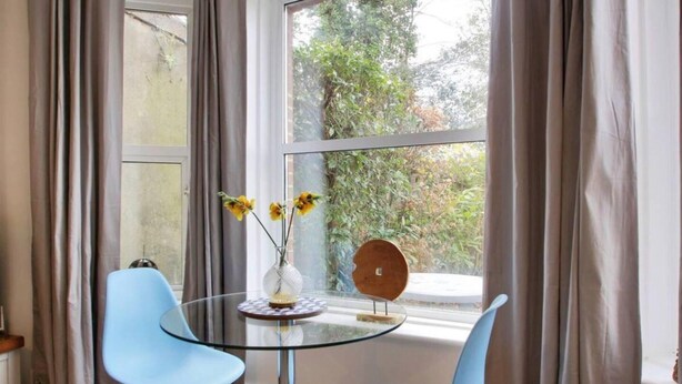 A room with a glass table and two blue chairs, a vase with flowers, and a view of greenery outside. - Stylish 1-bed Apartment - Heart of Tunbridge Wells (Royal Tunbridge Wells)