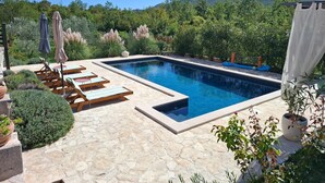 A rectangular swimming pool with a stone deck, surrounded by lounge chairs and umbrellas, set against a backdrop of lush greenery and mountains. - Villa Vinka (Vrgorac)