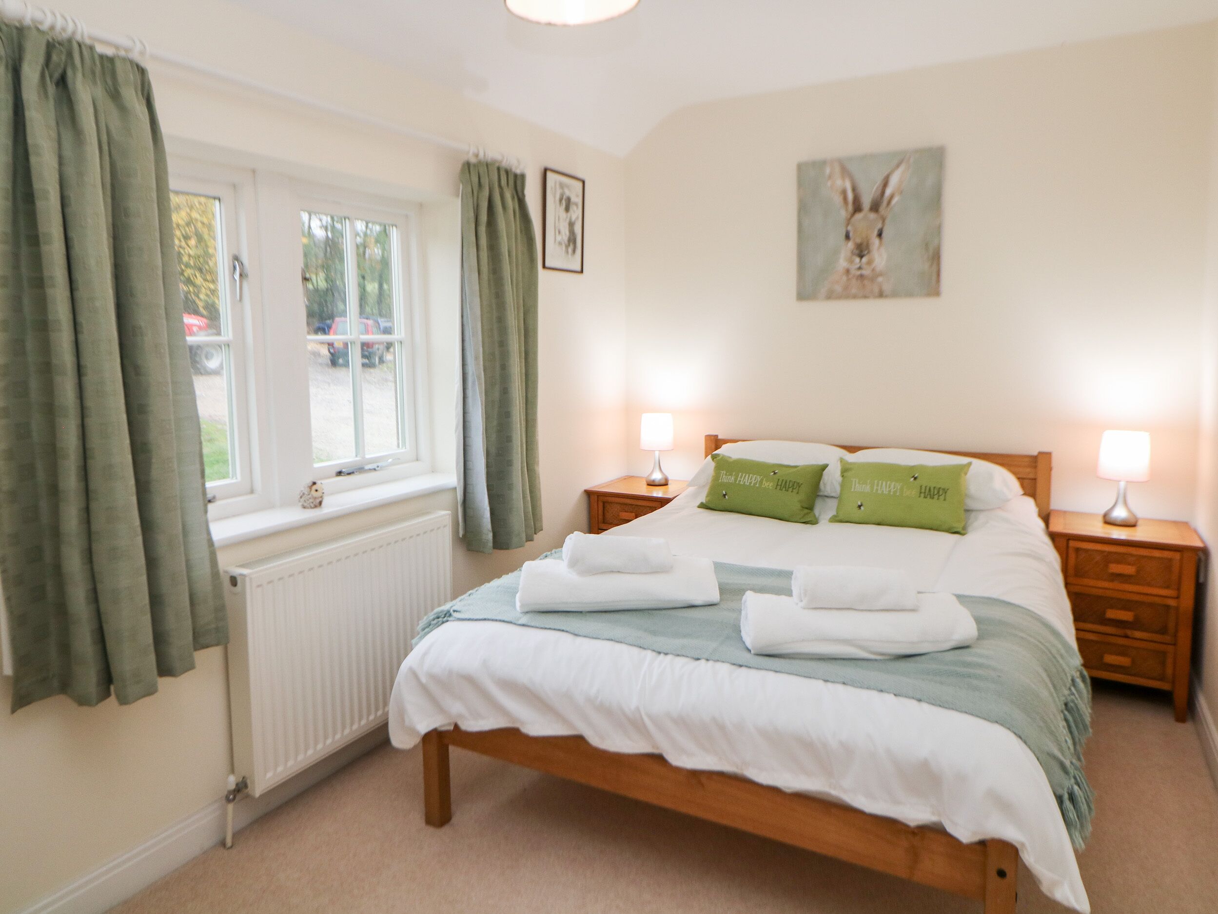 A neatly made bed with green pillows, a window with curtains, a radiator, and a painting of a rabbit on the wall.