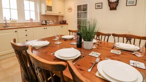 A dining room with a wooden table set for four, surrounded by wooden chairs. There is a kitchen in the background with white cabinets and a window. - Lower Cowden Farm (Bakewell)