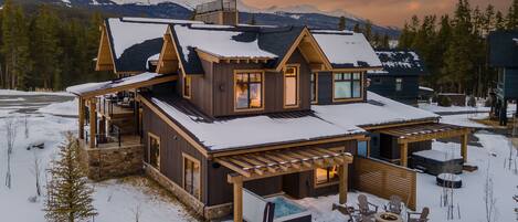 A large, multi-level wooden cabin with a snow-covered roof, a hot tub, and outdoor seating.