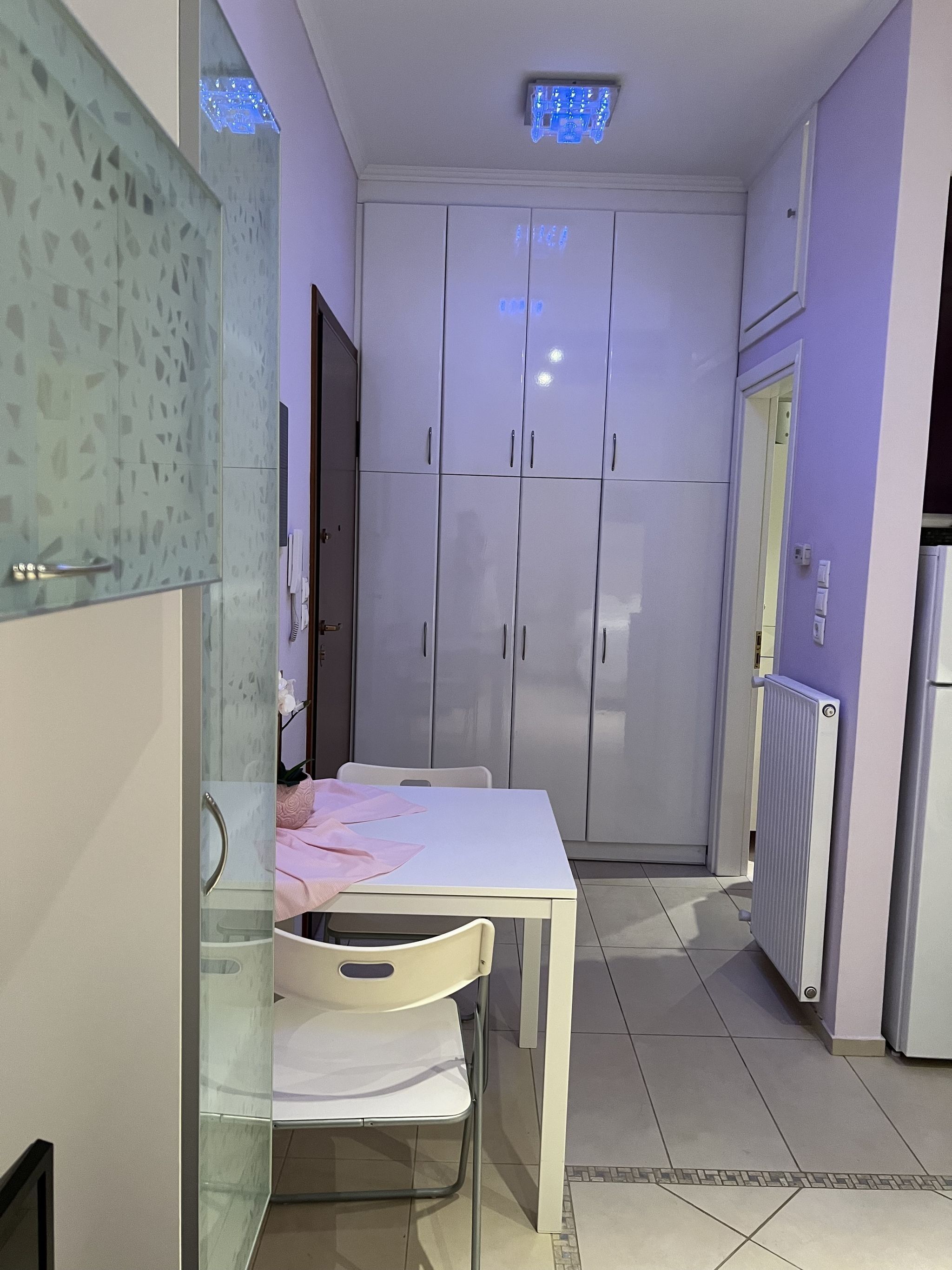 A narrow kitchen with a white table and chairs, a glass cabinet, and a white wardrobe.