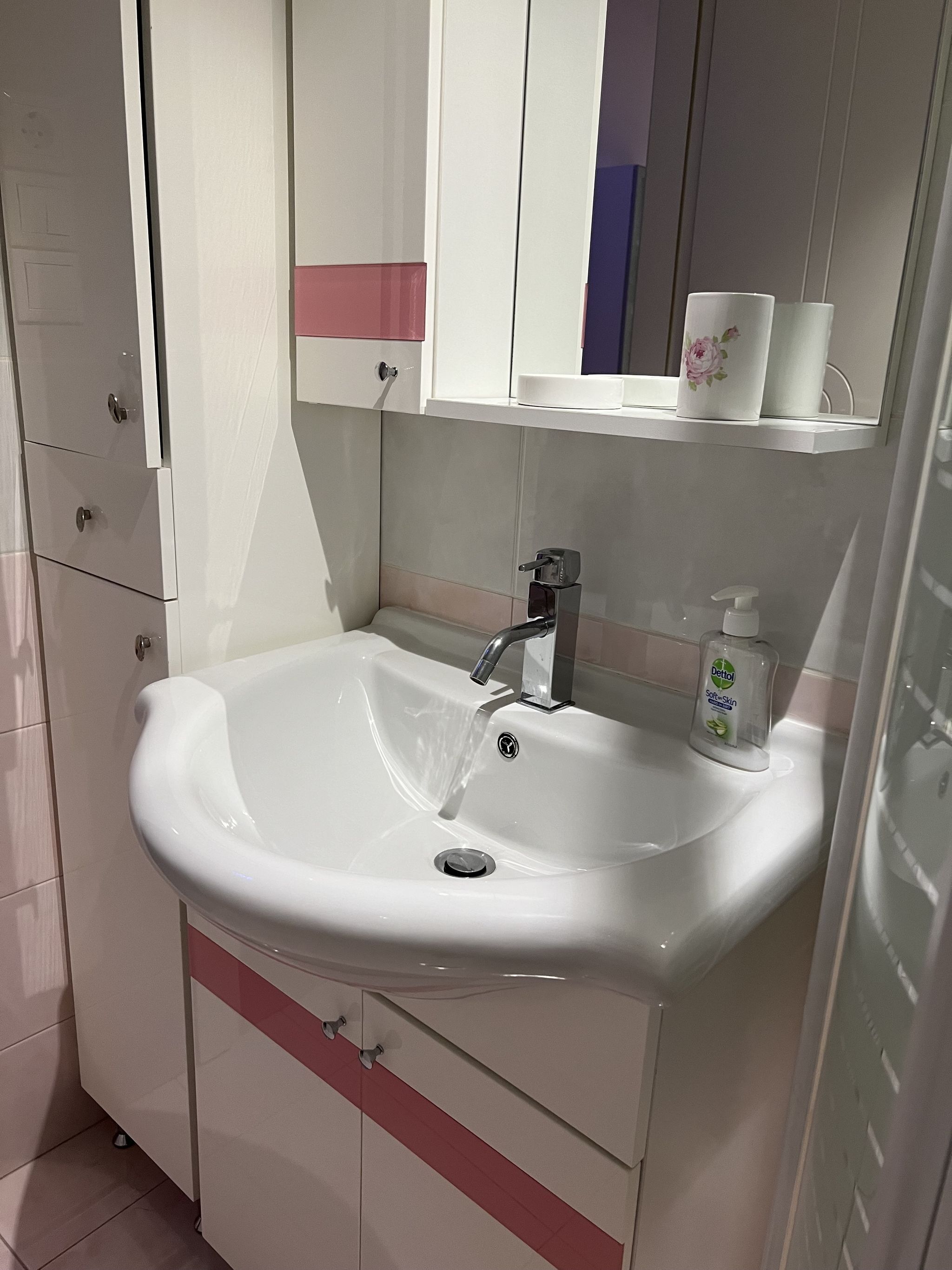 Un baño con un lavamanos blanco, un espejo y un estante con artículos de aseo.