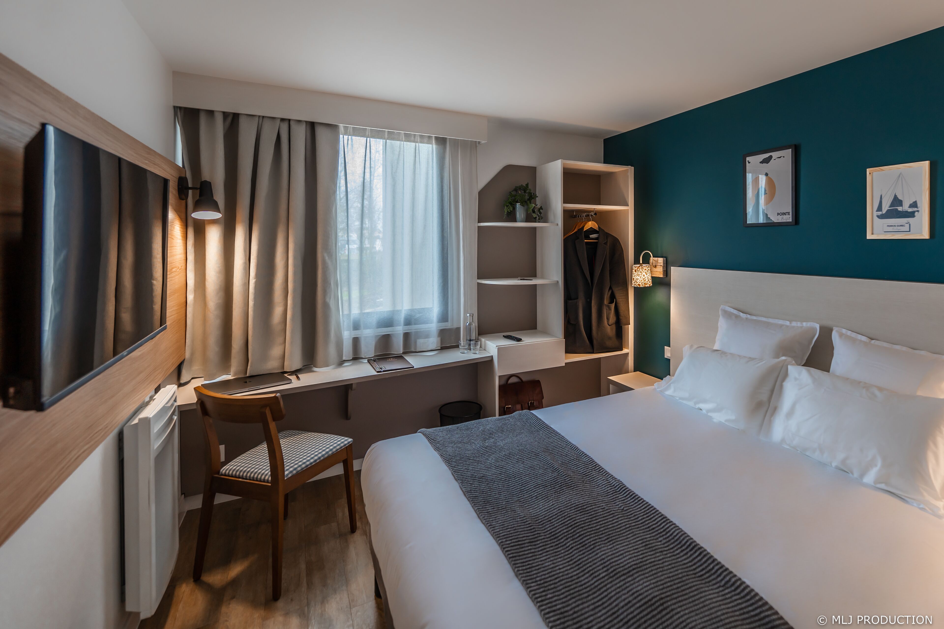 A modern hotel room with a large bed, a desk with a chair, a wardrobe, and a TV mounted on the wall.
