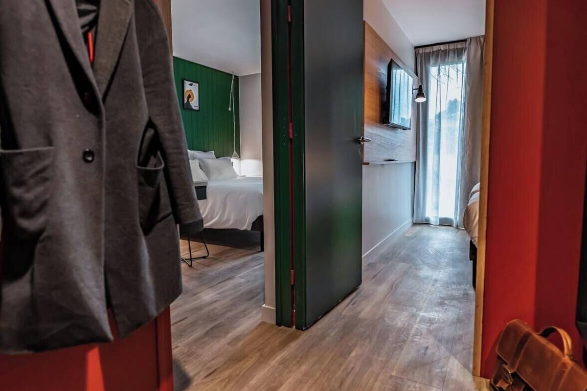 Ein Hotelkorridor mit Holzboden, Kleiderhaken, einem Bett an einer grünen Wand und einem Fenster mit Vorhängen.