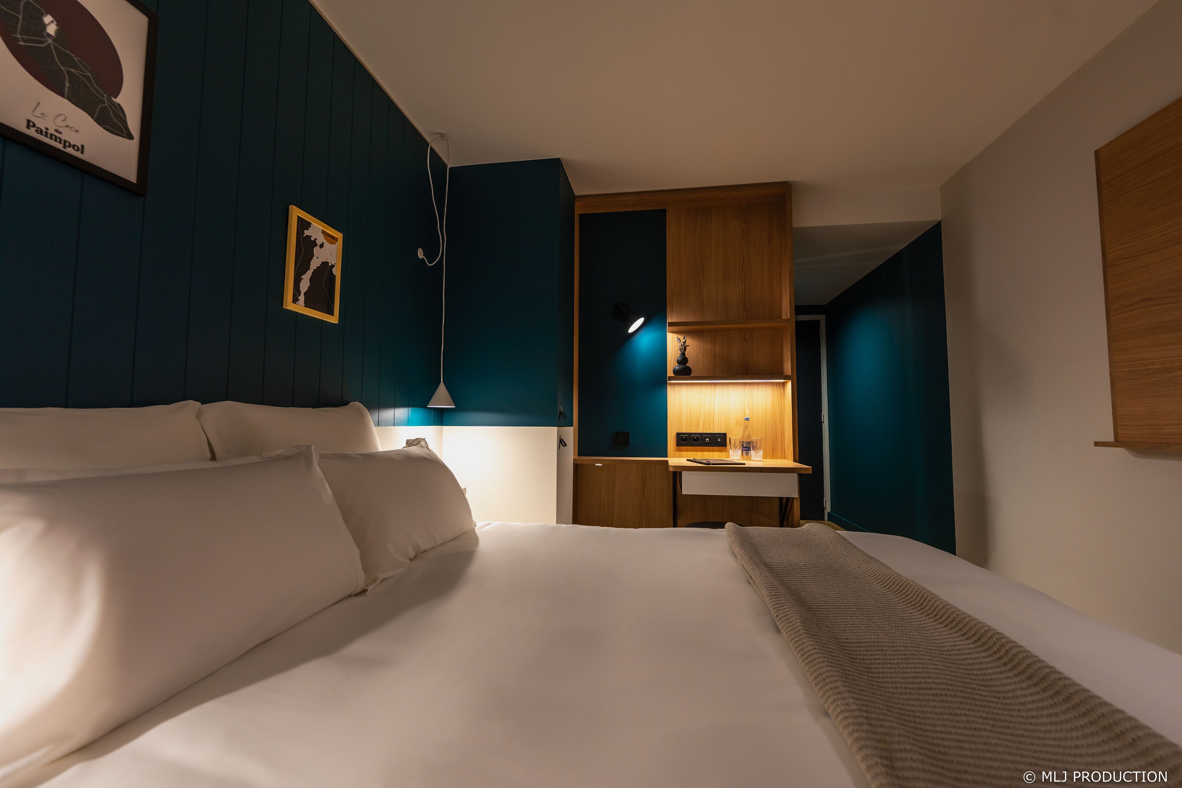 A hotel room with a large bed, a desk, and a chair. There is a lamp on the desk and a picture on the wall.