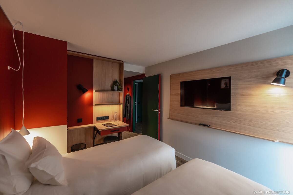 Ein modernes Hotelzimmer mit Bett, Nachttisch und Schreibtisch mit Stuhl. Ein Fernseher ist an der Wand befestigt und auf dem Schreibtisch steht eine Lampe.