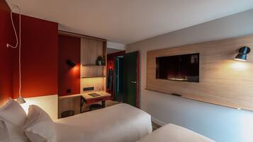 A modern hotel room with a bed, bedside table, and a desk with a chair. There is a TV mounted on the wall and a lamp on the desk.