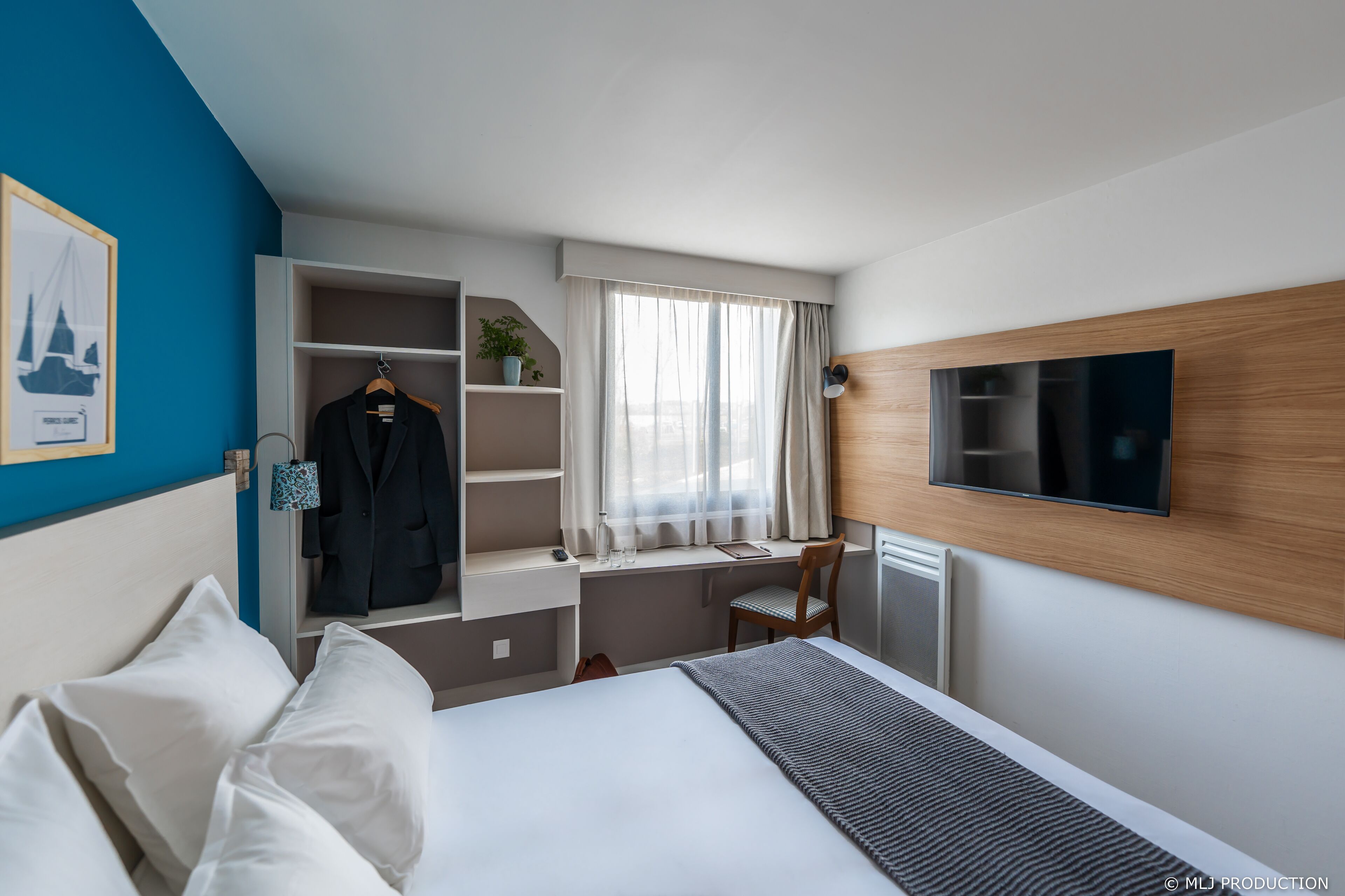A hotel room with a bed, a desk with a chair, a TV, a wardrobe, and a window with curtains.