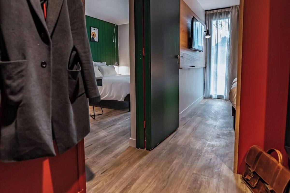 Ein Hotelkorridor mit Holzboden, Kleiderhaken, einem Bett an einer grünen Wand und einem Fenster mit Vorhängen.