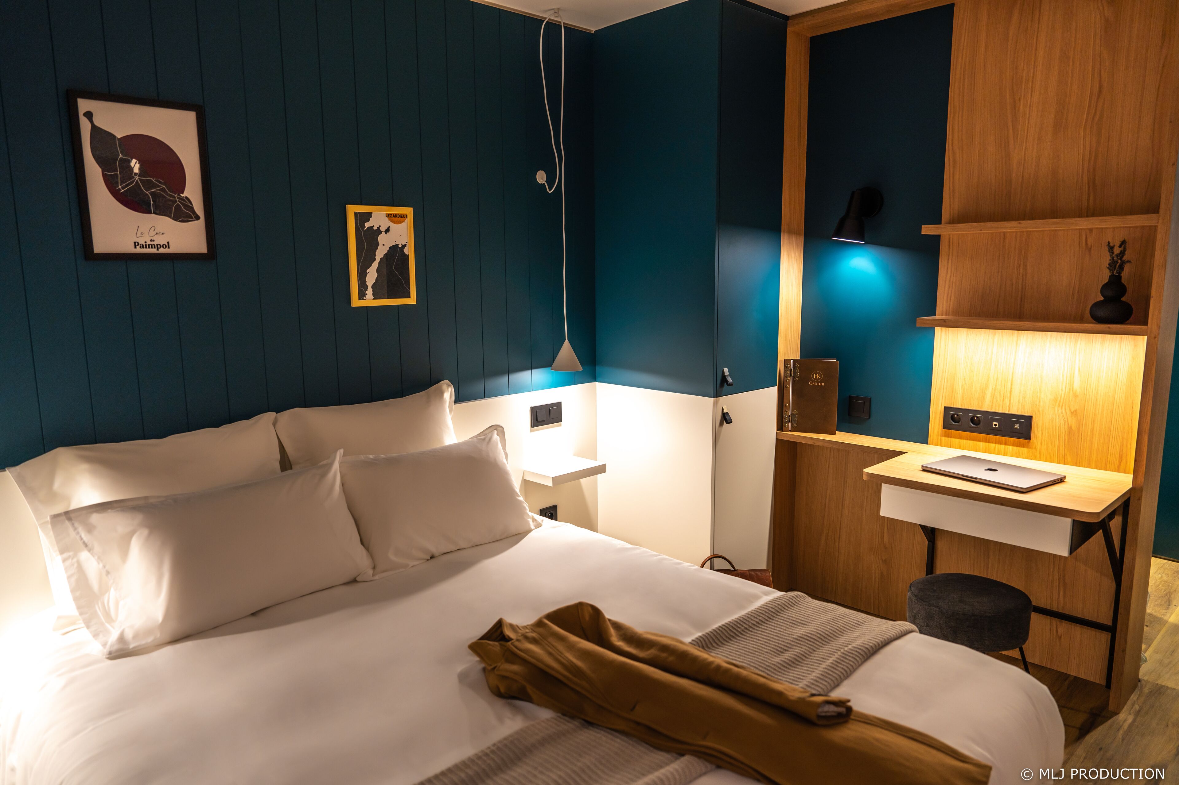 A hotel room with a bed, bedside tables, a desk with a laptop, a chair, and wall art.