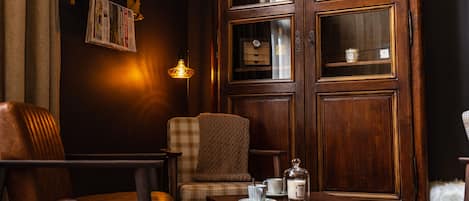 A cozy room with a wooden cabinet, a plaid armchair, a small table with a candle and a teapot, and a lamp.