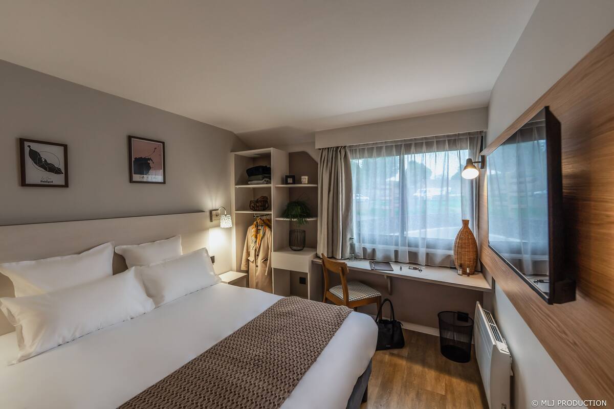 Ein Hotelzimmer mit einem großen Bett, einem Schreibtisch mit Stuhl, einem Fenster mit Vorhängen und einem an der Wand befestigten Fernseher.