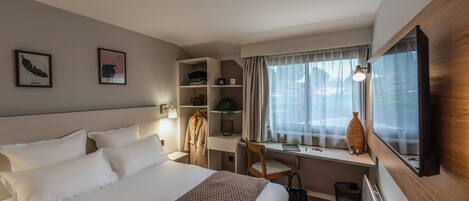 Habitació d'hotel amb un llit gran, un escriptori amb una cadira, una finestra amb cortines i un televisor penjat a la paret.