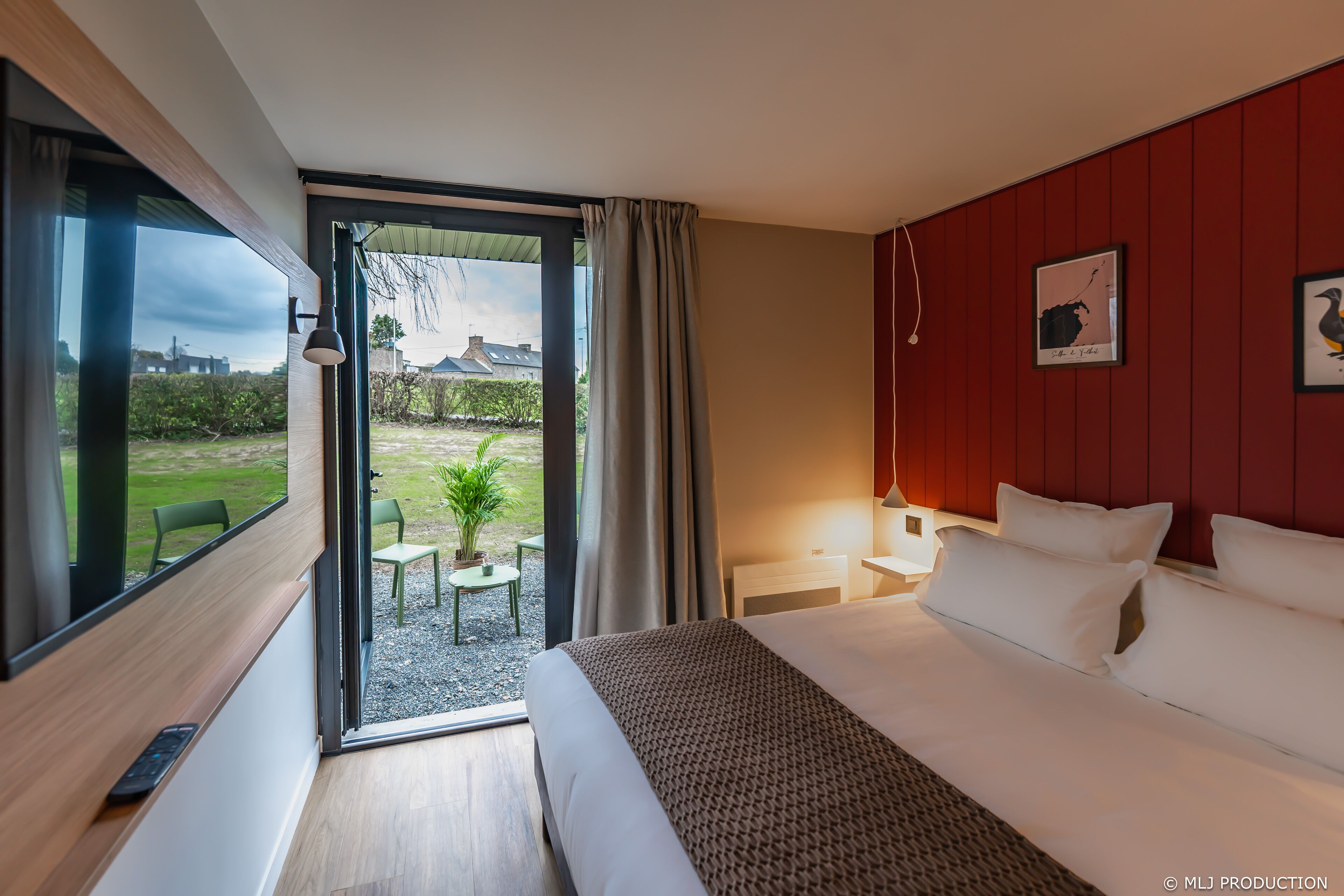 A modern hotel room with a large bed, a desk, and a view of the garden through the door.