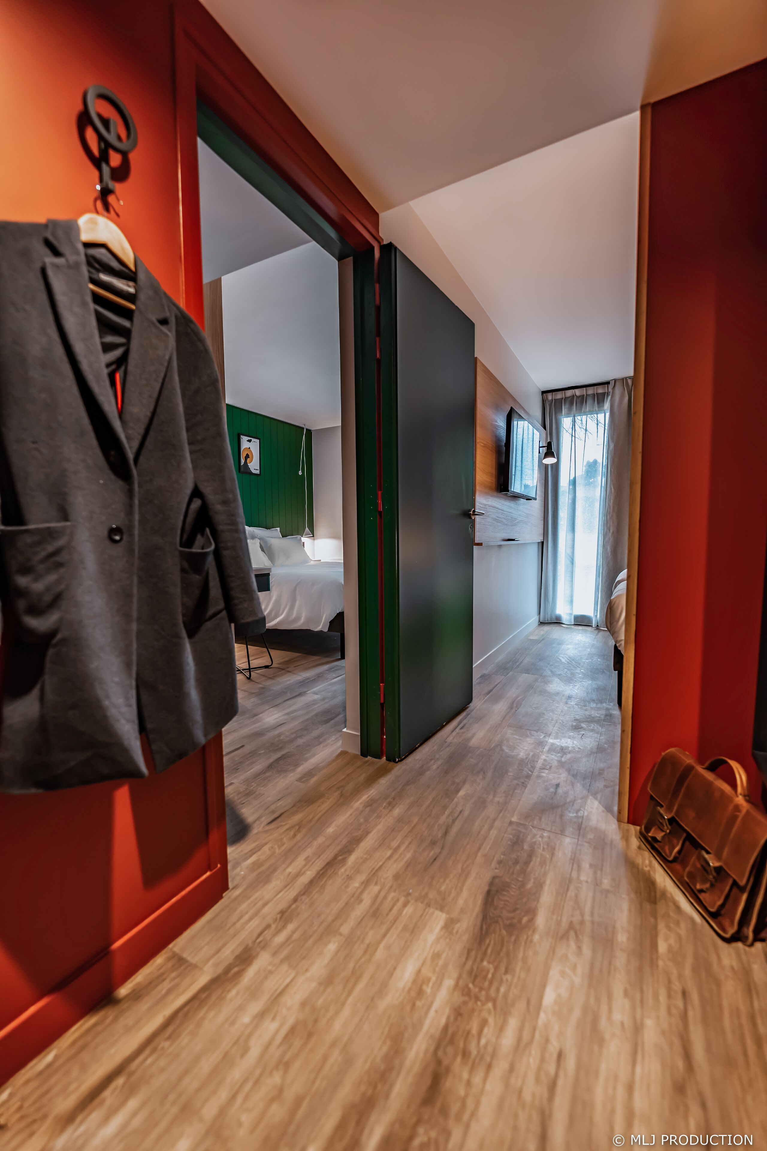 Ein Hotelgang mit Holzboden, einem Kleiderständer mit Jacke, einem Bett mit weißer Bettwäsche und einem Koffer auf dem Boden.