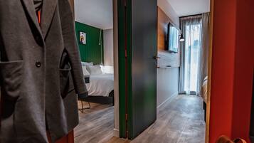 Ein Hotelgang mit Holzboden, einem Kleiderständer mit Jacke, einem Bett mit weißer Bettwäsche und einem Koffer auf dem Boden.