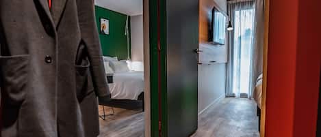 A hotel corridor with a wooden floor, a coat rack with a jacket, a bed with white bedding, and a suitcase on the floor.