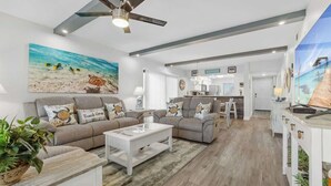 A spacious living room with a ceiling fan, a large painting of a beach scene, and a kitchen area in the background.