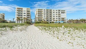 A beachfront hotel with multiple floors, a sandy pathway leading to the entrance, and a clear blue sky.