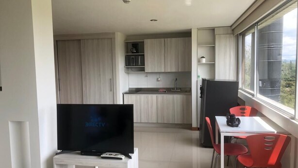Une cuisine moderne avec des meubles en bois, un coin repas avec des chaises rouges et un téléviseur posé sur un meuble blanc.