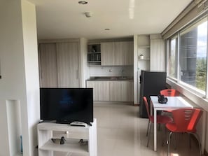 A modern kitchen with wooden cabinets, a dining area with red chairs, and a television on a white stand.