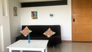 Une pièce avec une table blanche, un canapé sombre agrémenté de coussins à motifs, un tableau coloré, un petit appareil électronique fixé au mur et une porte en bois.