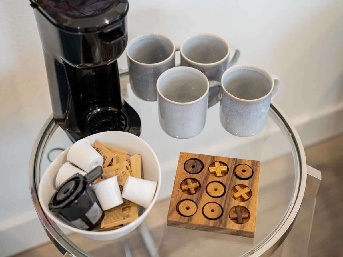 Una cafetera, seis tazas, un bol con aperitivos y una tabla de madera con símbolos sobre una mesa de vidrio.