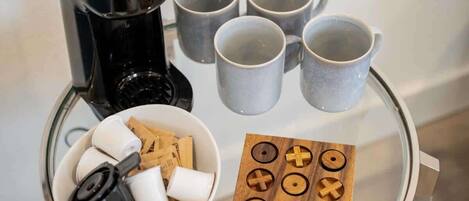 Una cafetera, seis tazas, un bol con aperitivos y una tabla de madera con símbolos sobre una mesa de vidrio.