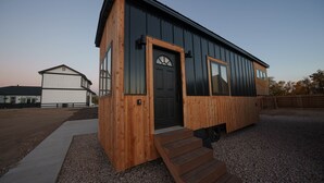 Eine kleine Holzhütte mit einer schwarzen Tür und Fenstern, einer Schotterauffahrt und einem Holzzaun im Hintergrund.