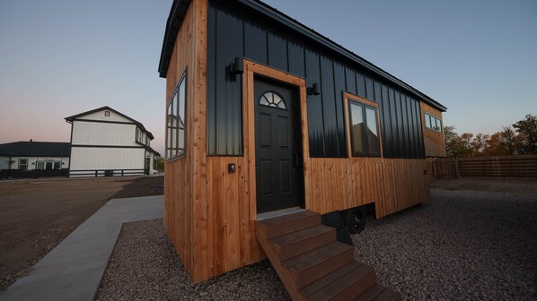 Une petite cabane en bois avec une porte et des fenêtres noires, une allée en gravier et une clôture en bois en arrière-plan.