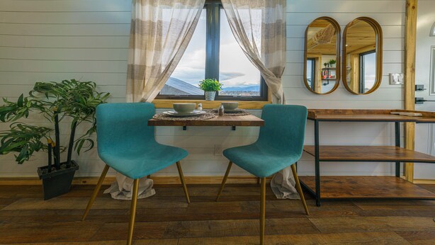 Ein Zimmer mit Fenster, zwei Türkis-Stühlen, einem Holztisch, einer Topfpflanze und einem Spiegel.