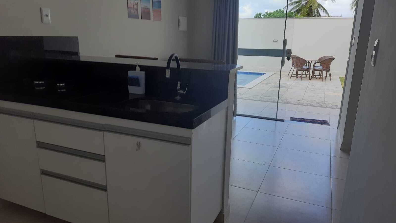 A modern kitchen with a black countertop and white cabinets, leading to an outdoor patio with a pool and chairs.