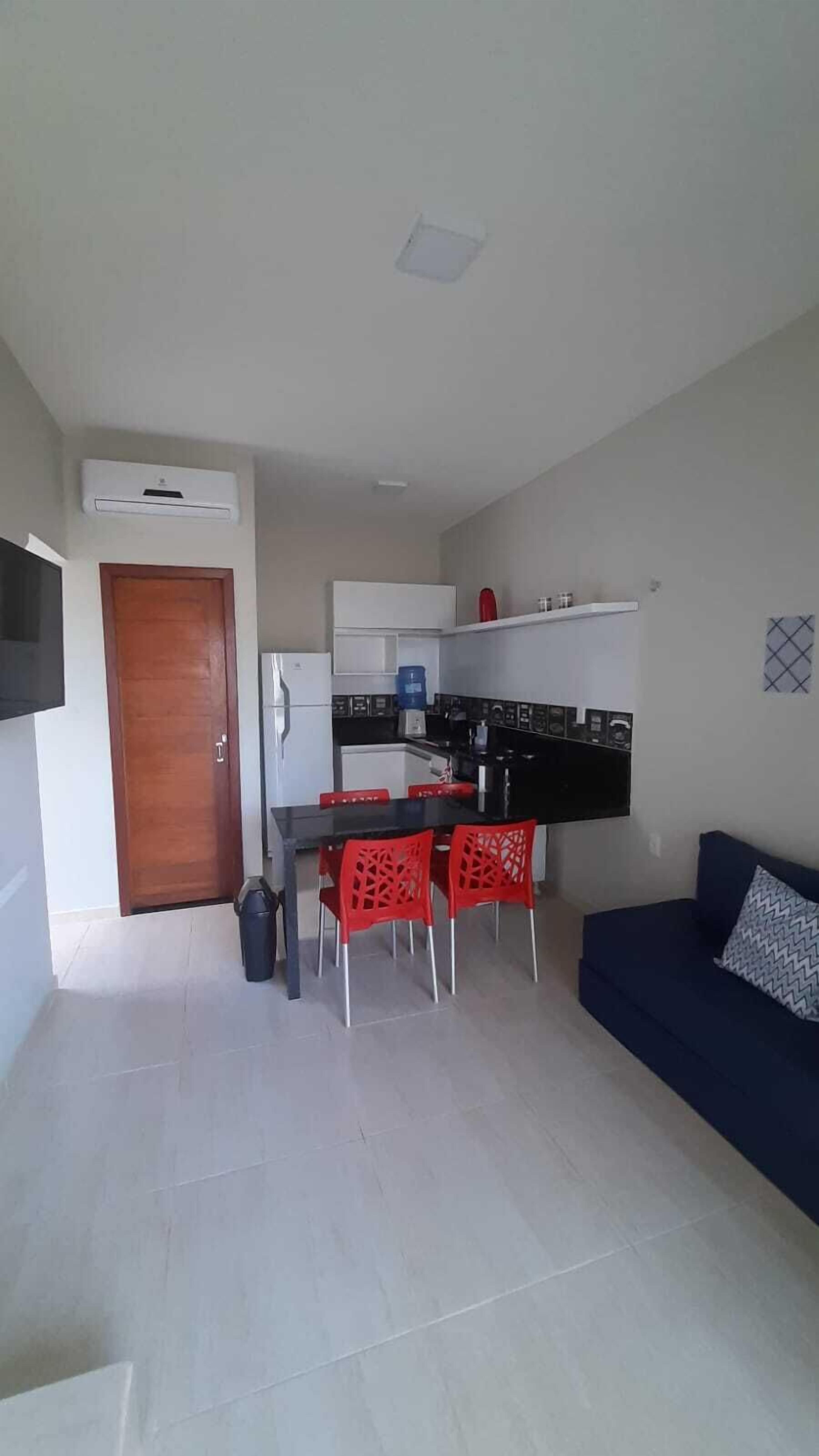 Een knus woongedeelte met een kitchenette, een eethoek met rode stoelen, een bank en een aan de muur bevestigde televisie.