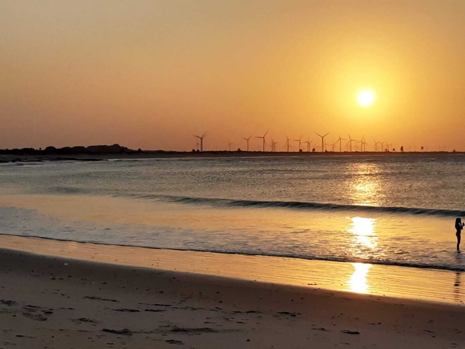 A beach with a sunset, waves, and wind turbines in the distance.