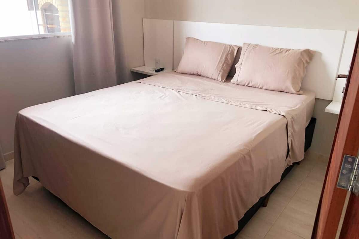 Un lit bien fait, avec une tête de lit blanche, une table de chevet sur laquelle se trouve une télécommande et une fenêtre avec des rideaux.