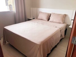 Una cama bien hecha con cabecera blanca, una mesita de noche con control remoto y una ventana con cortinas.